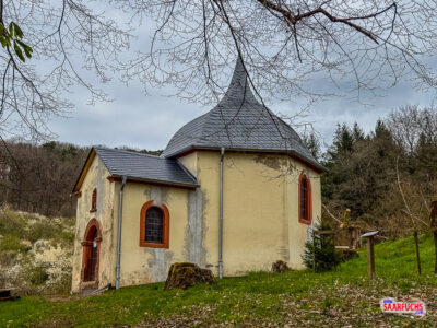 Historische Kreuzkapelle mit steinernem Mauerwerk inmitten einer Anlage mit verschiedenen Baumarten