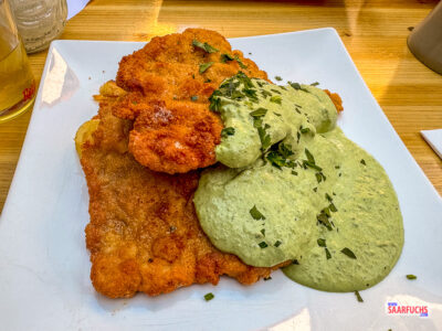 Ein paniertes Schnitzel mit goldbraunen Bratkartoffeln und einer großzügigen Portion grüner Kräutersauce auf einem weißen Teller