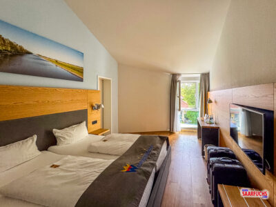 Innenansicht des geräumigen Hotelzimmers mit großen Betten und Blick durch das Fenster in den grünen Garten